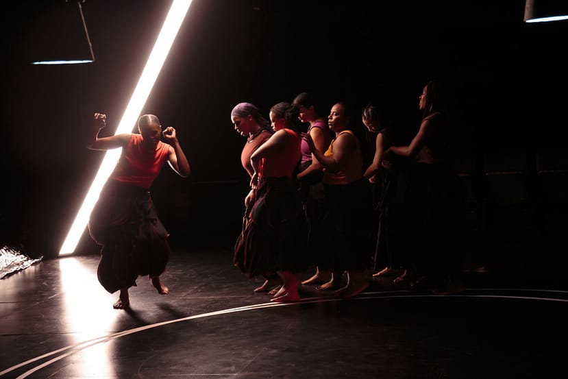six dancers in a bunch face another dancer with her arms up and hands in fist