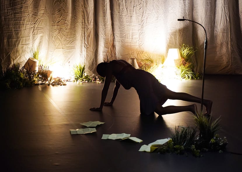 solo dancer on hands and knees with papers and florals scattered around her, lightly dimmed